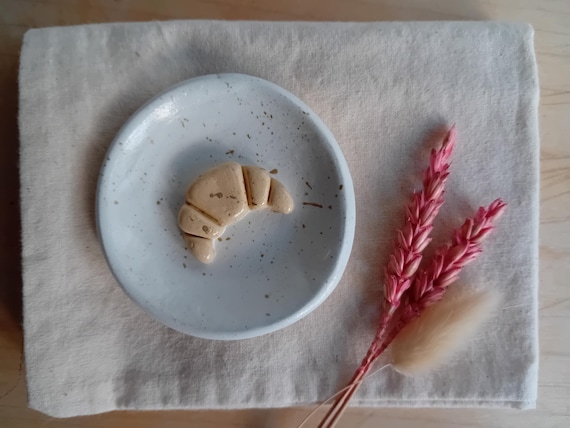 A tiny hand-sculpted clay dish shaped like a croissant with gold speckles — too cute and specific to be mass-produced, and perfect for corralling rings or earrings on a nightstand.
