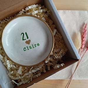 May include: A white ceramic trinket dish with a gold rim, inscribed with the number "21", a heart, and the name "Claire" in green. The dish is nestled in a cardboard box with shredded paper, accompanied by dried flowers.