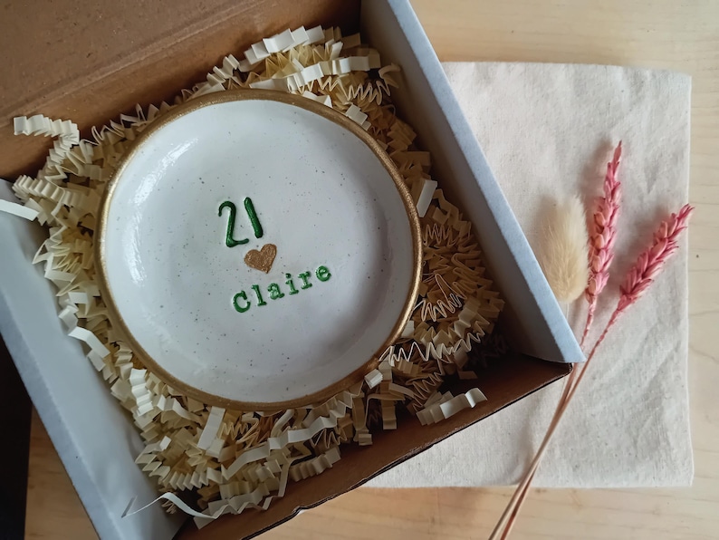 May include: A white ceramic trinket dish with a gold rim, inscribed with the number "21", a heart, and the name "Claire" in green. The dish is nestled in a cardboard box with shredded paper, accompanied by dried flowers.