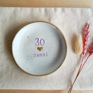 May include: A white ceramic dish with a gold rim, featuring the number "30", a heart, and the name "Sammi" in purple. The dish rests on a beige cloth, with pink and tan dried flowers to the side.