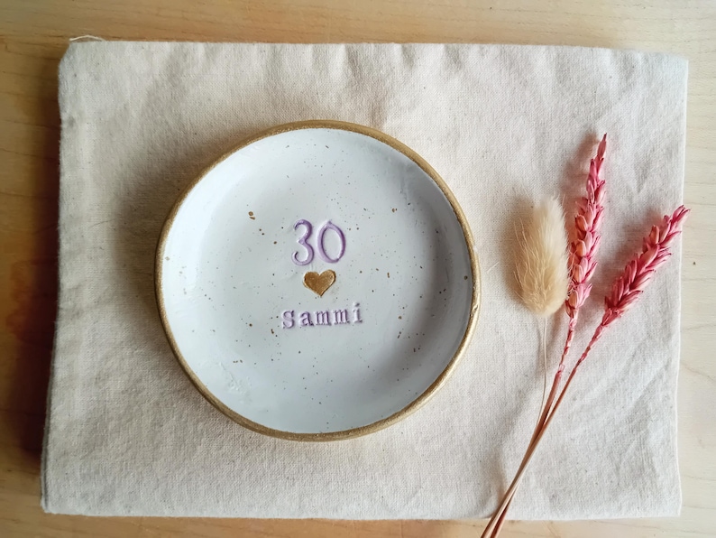 May include: A white ceramic dish with a gold rim, featuring the number "30", a heart, and the name "Sammi" in purple. The dish rests on a beige cloth, with pink and tan dried flowers to the side.