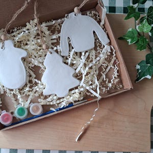 May include: A craft kit containing three white ceramic ornaments shaped like a mitten, a Christmas tree, and a jumper. Each ornament has a jute string and a wooden bead. The kit includes three small paint pots in red, green, and tan, and a paintbrush.