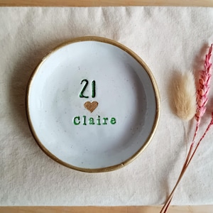 May include: A white ceramic ring dish with a gold rim, featuring the number "21", a heart, and the name "Claire" in green lettering. The dish is on a beige linen surface, with dried pink flowers and a beige fluffy plant.