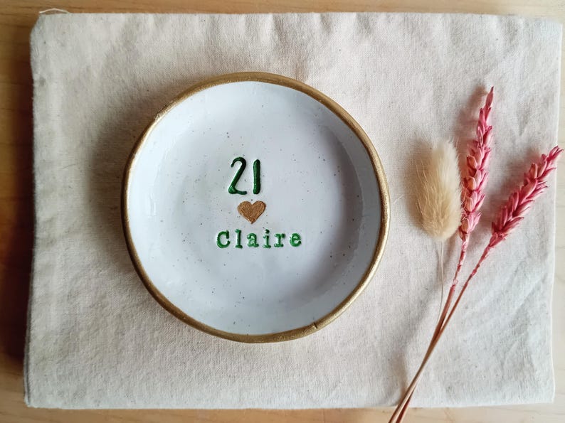 May include: A white ceramic ring dish with a gold rim, featuring the number "21", a heart, and the name "Claire" in green lettering. The dish is on a beige linen surface, with dried pink flowers and a beige fluffy plant.