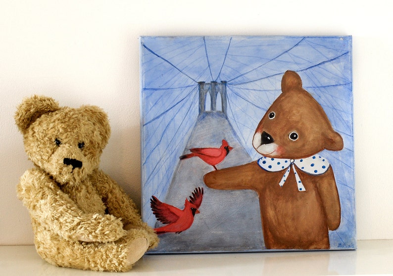 May include: A plush teddy bear sits next to a square painting. The painting depicts a brown bear with a blue and white polka-dot collar, pointing at two red cardinals. The background features a blue sky and a bridge.