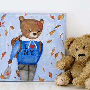 May include: A whimsical painting of a bear wearing a blue sweater with "I ❤️ NY" and a backpack, surrounded by falling leaves. A plush teddy bear sits beside the artwork. The painting is on a square canvas.