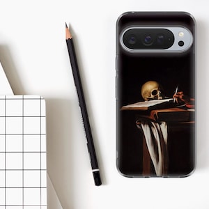 May include: A clear phone case featuring a painting of a skull on a table with a book and a hand holding a pen. A black pencil and a white notebook with a black grid pattern are also visible.