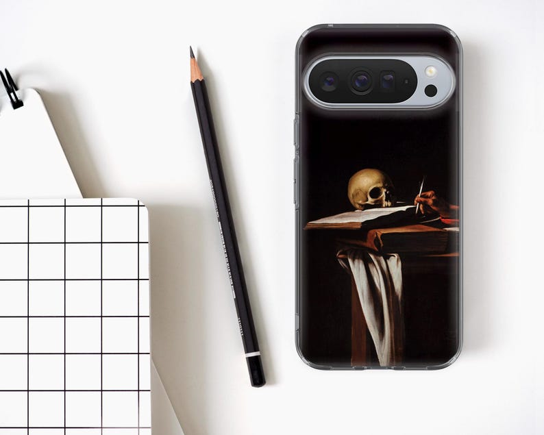May include: A clear phone case featuring a painting of a skull on a table with a book and a hand holding a pen. A black pencil and a white notebook with a black grid pattern are also visible.