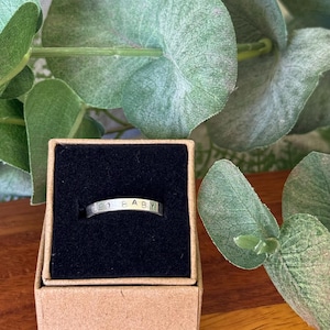 May include: A silver ring with the words "LEO BABY" engraved on it, presented in a small, open, brown cardboard box with a black interior. The ring is centered in the box. Green leafy foliage is in the background.