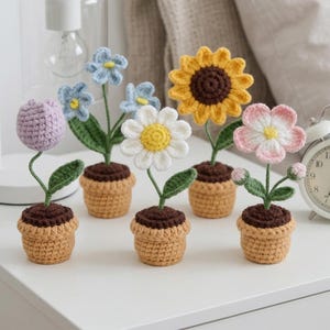 Puede incluir: Una colección de flores de ganchillo hechas a mano en macetas pequeñas. Las flores incluyen un tulipán morado, nomeolvides azules, una margarita blanca, un girasol amarillo y una flor rosa. Cada flor tiene hojas verdes y está en una maceta beige con tierra marrón.