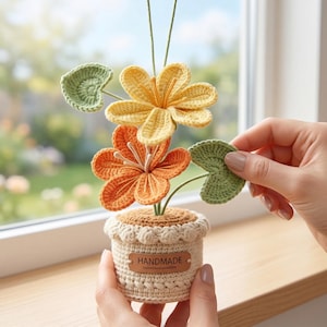 Könnte beinhalten: Ein handgefertigtes Häkelblumenarrangement in einem kleinen Topf. Das Arrangement zeigt gelbe und orangefarbene Blüten mit grünen Blättern. Der Topf ist beige mit einem gewellten Rand und einem "HANDMADE"-Etikett.