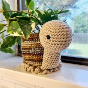 May include: A handmade crochet snail planter in beige with black eyes, holding a green and white plant. The snail's shell is a mix of brown, blue, and tan yarn. The planter sits on a white surface near a window.