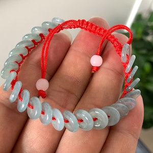 以下が含まれることがあります： 薄い緑色の翡翠のビーズと赤い紐の留め具が付いた翡翠のブレスレット。ブレスレットには、紐にピンクのビーズが2つ付いています。