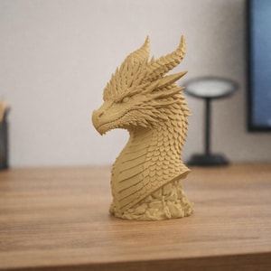 Puede incluir: Una escultura detallada de la cabeza de un dragón de color beige. El dragón tiene escamas texturizadas, cuernos y una cara detallada. La escultura está sobre un escritorio de madera, con un fondo borroso que incluye un monitor de computadora y un portalápices.