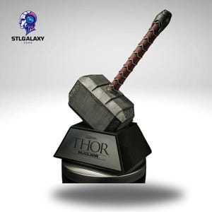 May include: A silver replica of Thor's hammer, Mjolnir, with a brown leather handle. The hammer is on a black base with the words "MARVEL STUDIOS THOR MJOLNIR" inscribed on it.