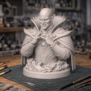 Puede incluir: Busto de resina sin pintar de un personaje calvo y sonriente con expresión malvada. La figura lleva una armadura ornamentada y una capa con hombreras puntiagudas. El busto está sobre una base redonda, con materiales de arte en el fondo.