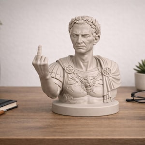 May include: A light gray bust of a Roman emperor, wearing a laurel wreath and military attire, giving the middle finger. The sculpture is on a wooden surface, with a notebook, pen, and glasses nearby.
