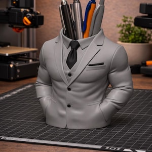 Puede incluir: Un portalápices gris con forma de traje de hombre, con corbata negra y bolígrafos en la zona de la cabeza. El portalápices tiene un diseño de traje de tres botones y está sobre una superficie de madera.
