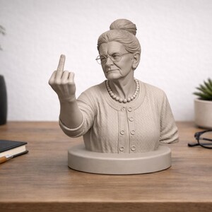 Könnte beinhalten: Eine hellgraue Büste einer älteren Frau mit Brille und Perlenkette, die ihren Mittelfinger zeigt. Die Skulptur steht auf einem runden Sockel, mit einem Cardigan mit Knöpfen. Der Hintergrund umfasst einen Schreibtisch, Pflanzen und eine Brille.