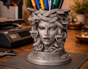 Peut inclure: Un porte-stylos en forme de tête de Méduse grise rempli de stylos et de crayons de différentes couleurs. La sculpture détaillée représente des serpents et une expression faciale sévère. Le support est posé sur une surface sombre, avec une lampe de bureau et d'autres objets en arrière-plan.
