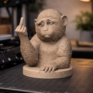 May include: A tan-colored monkey sculpture with a sad expression, giving the middle finger. The monkey is seated on a circular base. The sculpture is detailed and appears to be made of clay or a similar material.