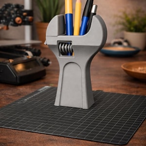 Puede incluir: Un organizador de escritorio gris en forma de llave inglesa que contiene bolígrafos y lápices. El organizador está hecho de un material sólido y se encuentra sobre una estera negra con un patrón de cuadrícula. Los bolígrafos y lápices son azules, amarillos y negros.