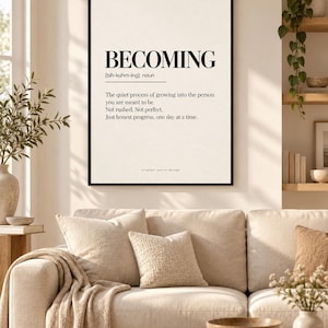 Puede incluir: Un cuadro decorativo enmarcado con la palabra BECOMING en letras mayúsculas negras, seguida de su pronunciación fonética y una definición que describe el proceso de convertirse en la persona que uno debe ser, un día a la vez.