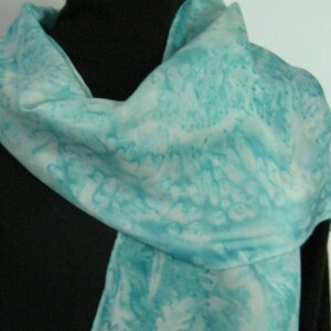 May include: A light blue scarf with a white and blue pattern. The scarf is draped over a black surface, showcasing its soft texture and color variations. The scarf's design features a watercolor-like effect.