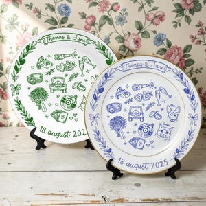 Puede incluir: Dos platos decorativos blancos con ilustraciones verdes y azules y bordes dorados. Los platos presentan dibujos temáticos de boda, los nombres "Thomas & Jane" y las fechas "18 agosto 202" y "18 agosto 2025".
