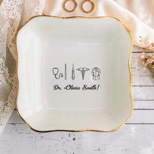 Könnte beinhalten: Weißes Keramik-Schmucktablett mit goldenem Rand, mit medizinischen Symbolen und dem Namen "Dr. Olivia Smith!" in schwarzer Schrift. Das Tablett ist quadratisch mit sanft geschwungenen Kanten, ideal für kleine Gegenstände.