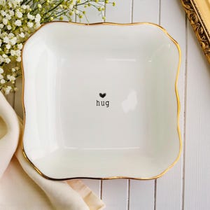 Könnte beinhalten: Ein weißer quadratischer Keramik-Teller mit gewelltem Goldrand. Der Teller hat ein schwarzes Herz und das Wort "hug". Der Teller steht auf einer weißen Holzoberfläche, mit Blumen und einem Tuch im Hintergrund.