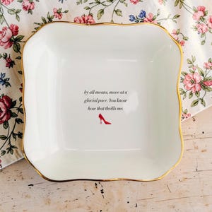 May include: White square ceramic dish with a gold trim. The dish has the text "by all means, move at a glacial pace. You know how that thrills me." and a red high-heeled shoe graphic.