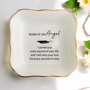 Könnte beinhalten: Eine weiße Keramik-Schmuckschale mit goldenem Rand. Die Schale trägt den Text "MAMA OF AN Angel" und eine Federgrafik. Darunter steht das Zitat: "I carried you every second of your life. And I will carry your love foe every second of mine."