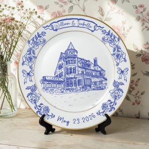 May include: White ceramic plate with a blue illustration of a house, floral border, and gold trim. The plate has the names "William & Isabella" and the date "May 23, 2026". The plate is displayed on a black stand.