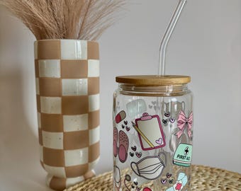 May include: A clear glass reusable water bottle with a bamboo lid and a straw. The bottle has a colorful design featuring medical symbols, a stethoscope, a bandage, a face mask, and the words "First Aid".