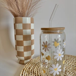 May include: A clear glass jar with a wooden lid and a glass straw. The jar is decorated with white daisies and yellow hearts. The jar is sitting on a woven straw mat.