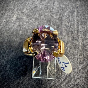 Może przedstawiać: Złoty pierścionek z dużym, owalnym, fasetowanym ametystem. Pierścionek znajduje się na stojaku z metką cenową z napisem "47."