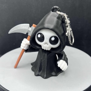 May include: A black and white 3D printed keychain featuring a cartoon Grim Reaper holding a scythe. The reaper has large eyes and a skull-like face.