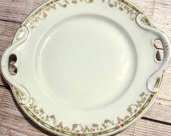May include: A white porcelain serving plate with a floral design in shades of pink and green. The plate has two handles and a gold trim.