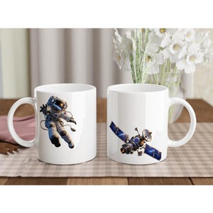 Pode incluir: Duas canecas de cerâmica branca com gráficos de temática espacial. Uma caneca exibe um astronauta com traje branco flutuando no espaço, enquanto a outra apresenta um satélite detalhado com painéis solares azuis e corpo metálico sobre fundo branco.