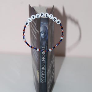 Könnte beinhalten: Ein blaues und rotes Perlen-Lesezeichen mit weißen Buchstabenperlen, die "WITCHLING" buchstabieren. Das Lesezeichen ist um ein Buch mit dem Titel "Throne of Glass" von Sarah J. Maas gewickelt.