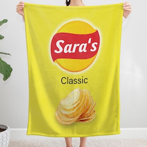 Peut inclure: Une couverture jaune avec un motif de paquet de chips. Le design comprend un logo rouge et jaune avec le texte "Sara's" et "Classic". Une pile de chips est également représentée.