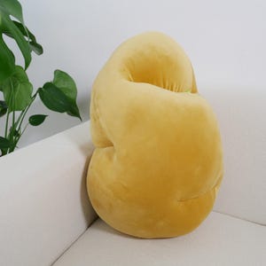 Puede incluir: Un cojín grande, amarillo y de felpa con forma de ocho. El cojín está sentado en un sofá blanco.