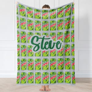 Può includere: Una coperta grigia con un motivo ripetuto di lattine di soda verdi e rosse. La parola "Steve" è scritta in verde al centro della coperta. La coperta è tenuta da una persona.