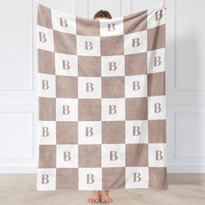 May include: A soft, plush blanket with a checkerboard pattern in brown and white. Each brown square features a white "B" in the centre. The blanket is held up, showcasing its design and texture. A cosy home decor item.