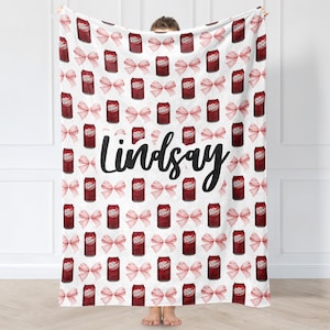 Könnte beinhalten: Weiße Decke mit einem sich wiederholenden Muster aus roten Dr. Pepper-Dosen und rosa Schleifen. Der Name "Lindsay" ist in schwarzer Schrift über die Mitte der Decke geschrieben.