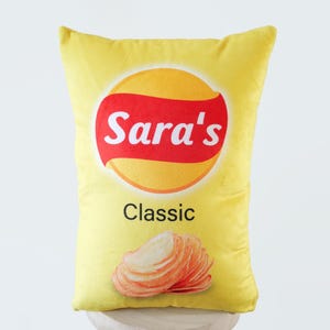 Könnte beinhalten: Gelbes Kissen mit einem roten und gelben Chipstüten-Design. Die Tüte trägt den Schriftzug "Sara's Classic" und zeigt ein Bild von Kartoffelchips.
