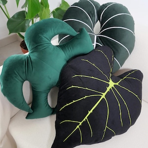 Puede incluir: Tres almohadas decorativas con forma de hojas. Una almohada es verde con rayas blancas, otra es verde oscuro y la última es negra con un patrón de vetas verde claro.