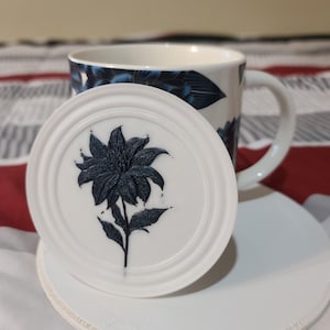 May include: A white ceramic coaster with a dark blue floral design, alongside a matching mug. The mug displays a similar blue floral pattern on a white backdrop. The coaster is circular and has a raised edge.