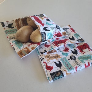 May include: Two white fabric reusable bags with a farm-themed print featuring chickens, pigs, and windmills. The bags are filled with three brown potatoes.